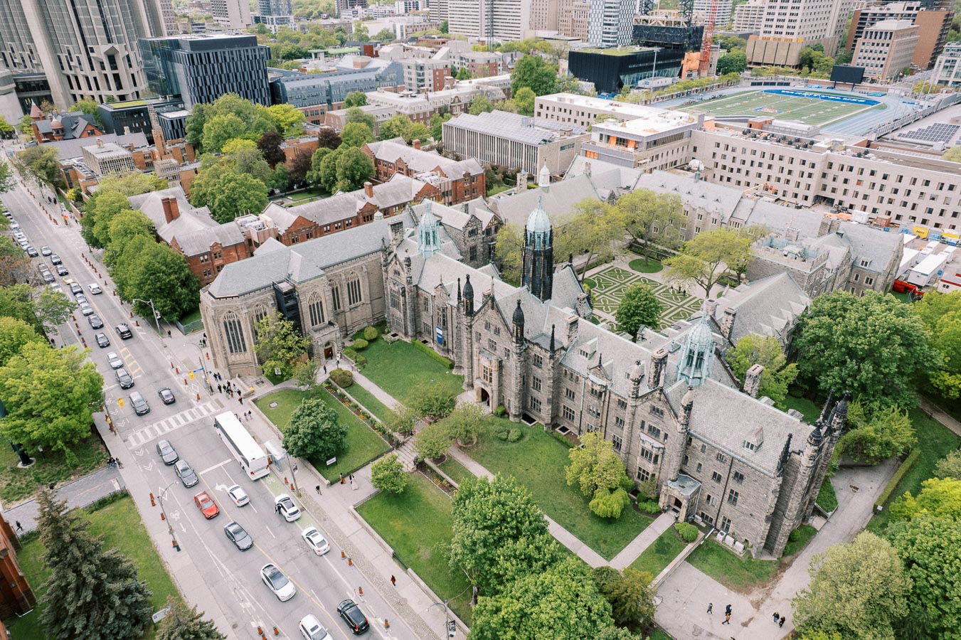 Aerial view of a historic university campus with gothic architecture, surrounded by lush green trees, a bustling city street, and modern buildings in the background.