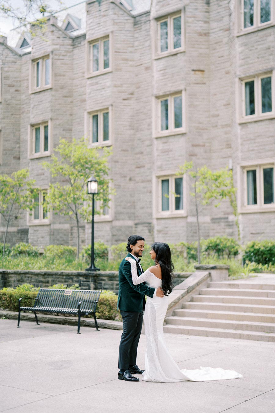 A couple in elegant wedding attire shares a joyful moment in front of a historic stone building, surrounded by greenery and steps leading to the entrance.