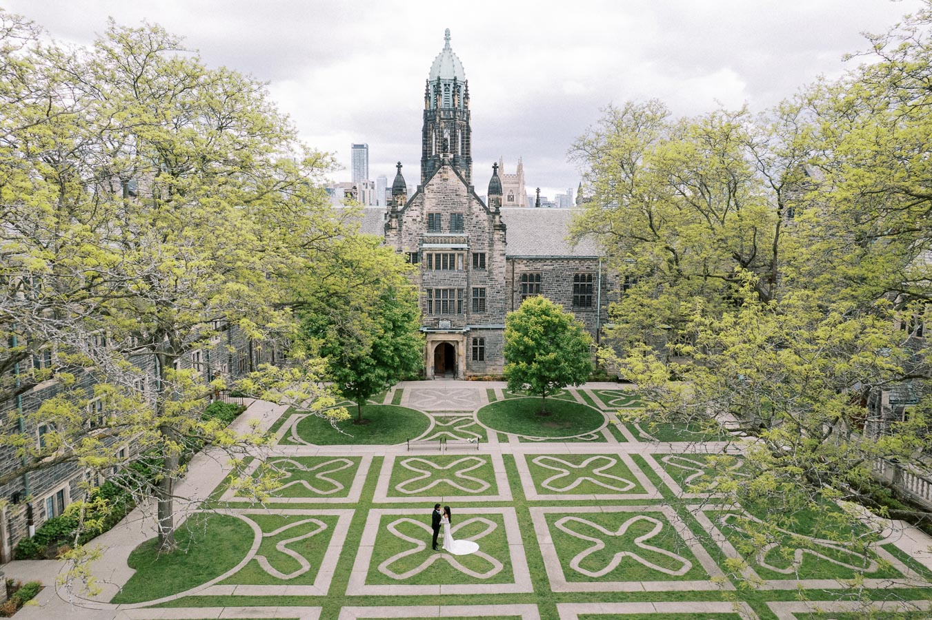 Aerial view of a historic, stone university building with intricate Gothic architecture, surrounded by lush green trees. The courtyard features geometric patterns on the grass, with a couple standing in the center, creating a picturesque scene.