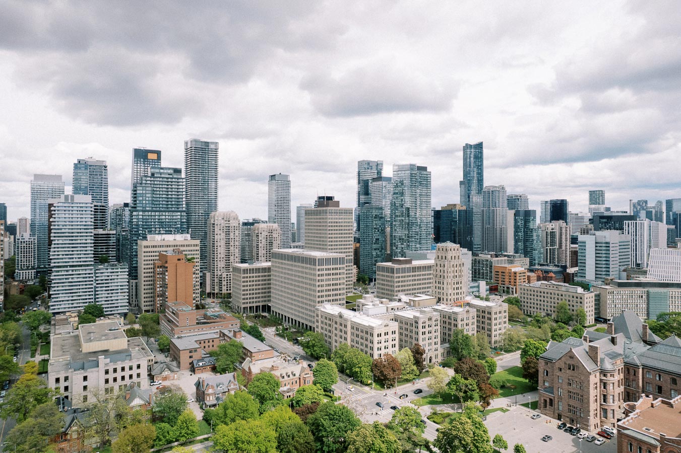 Aerial view of a modern city skyline with tall skyscrapers and lush green spaces under a cloudy sky, showcasing urban architecture and greenery.