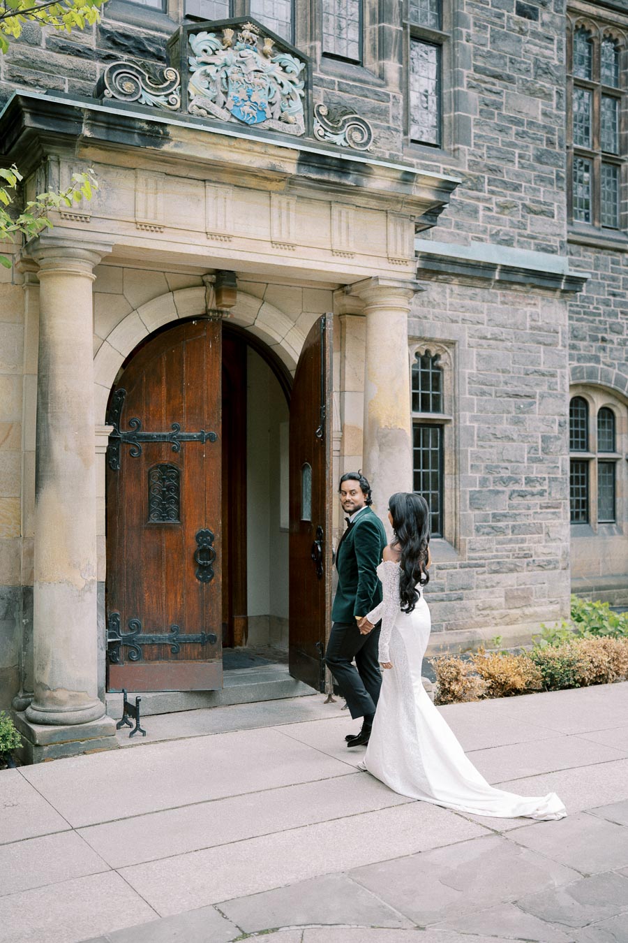 Couple entering a historic building with ornate wooden doors and stone architecture, dressed in formal attire.