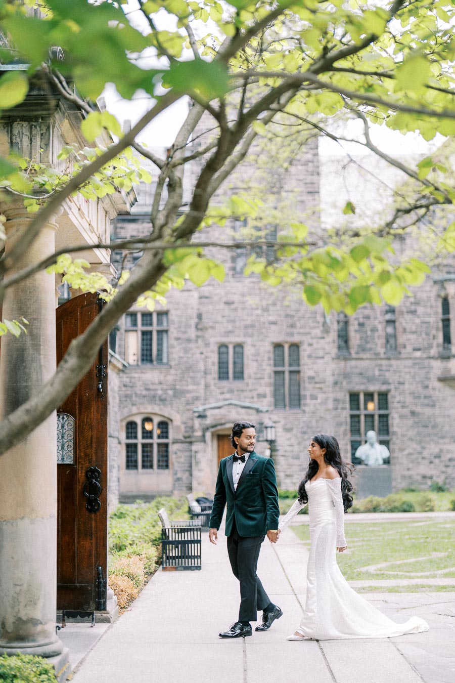 Elegant bride and groom walking hand in hand by historic stone building, surrounded by lush greenery and spring foliage, capturing romantic wedding moment