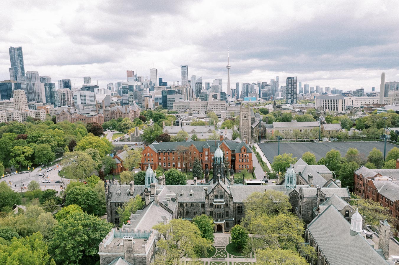 Aerial view of a historic university campus surrounded by lush greenery in the foreground, with a modern city skyline and iconic tower visible in the background under a cloudy sky.