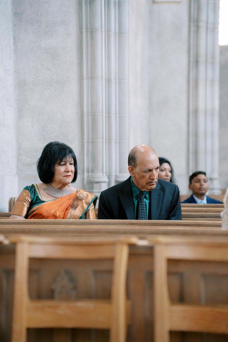Elderly couple sitting in a church pew during a ceremony, with the woman wearing a colorful saree and the man in a formal suit.
