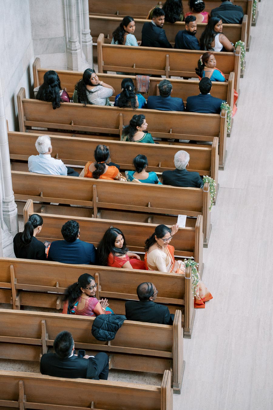 Aerial view of a diverse group of people seated in wooden church pews, dressed in colorful traditional attire, attentively looking towards the camera in a large, spacious hall decorated with floral arrangements.