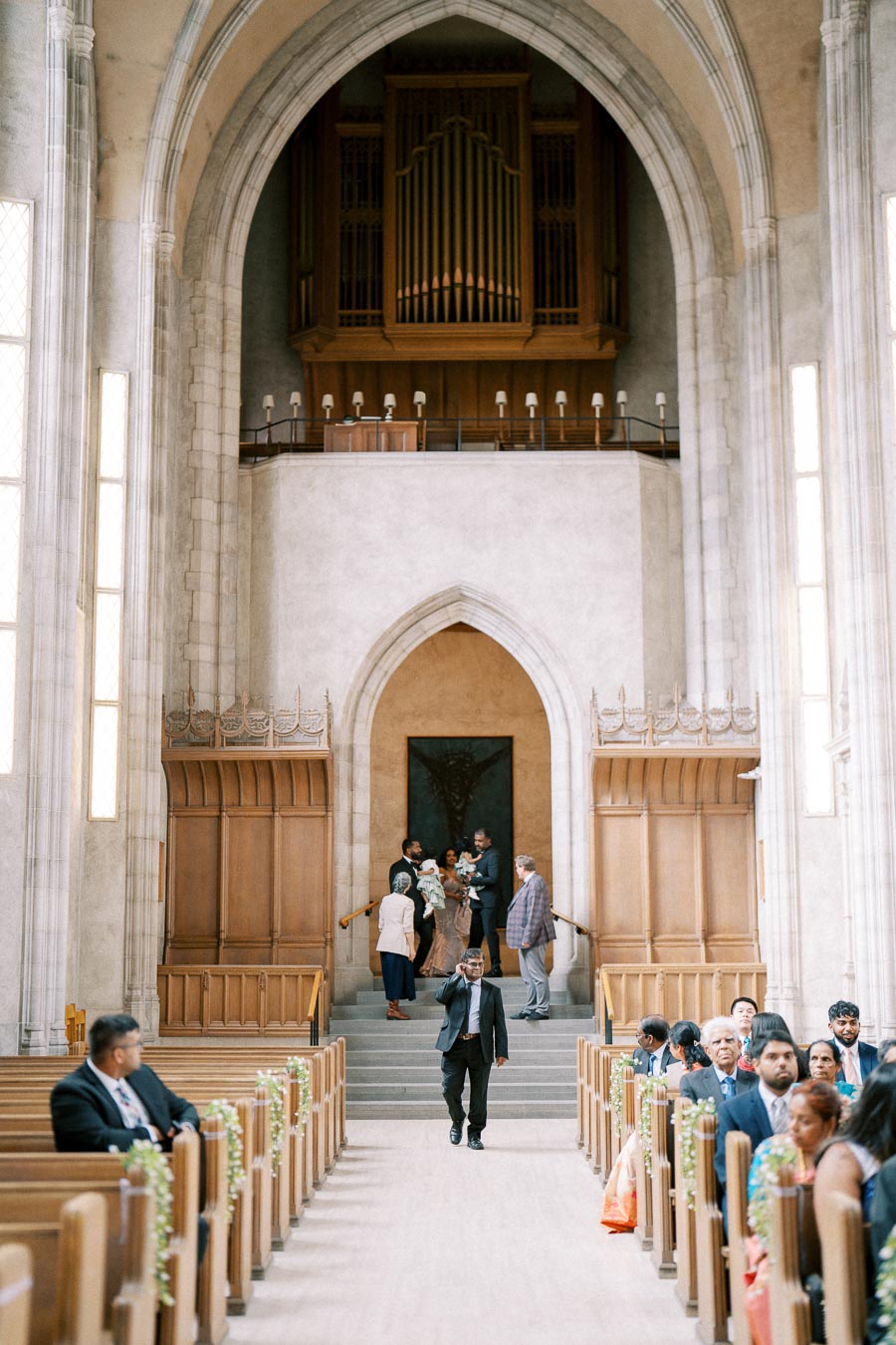 Elegant church interior with wooden pews, decorated for a wedding ceremony. A group of people are gathered near the altar, while a man in a suit walks down the aisle. Large organ pipes and stone archways add to the grandeur of the setting.