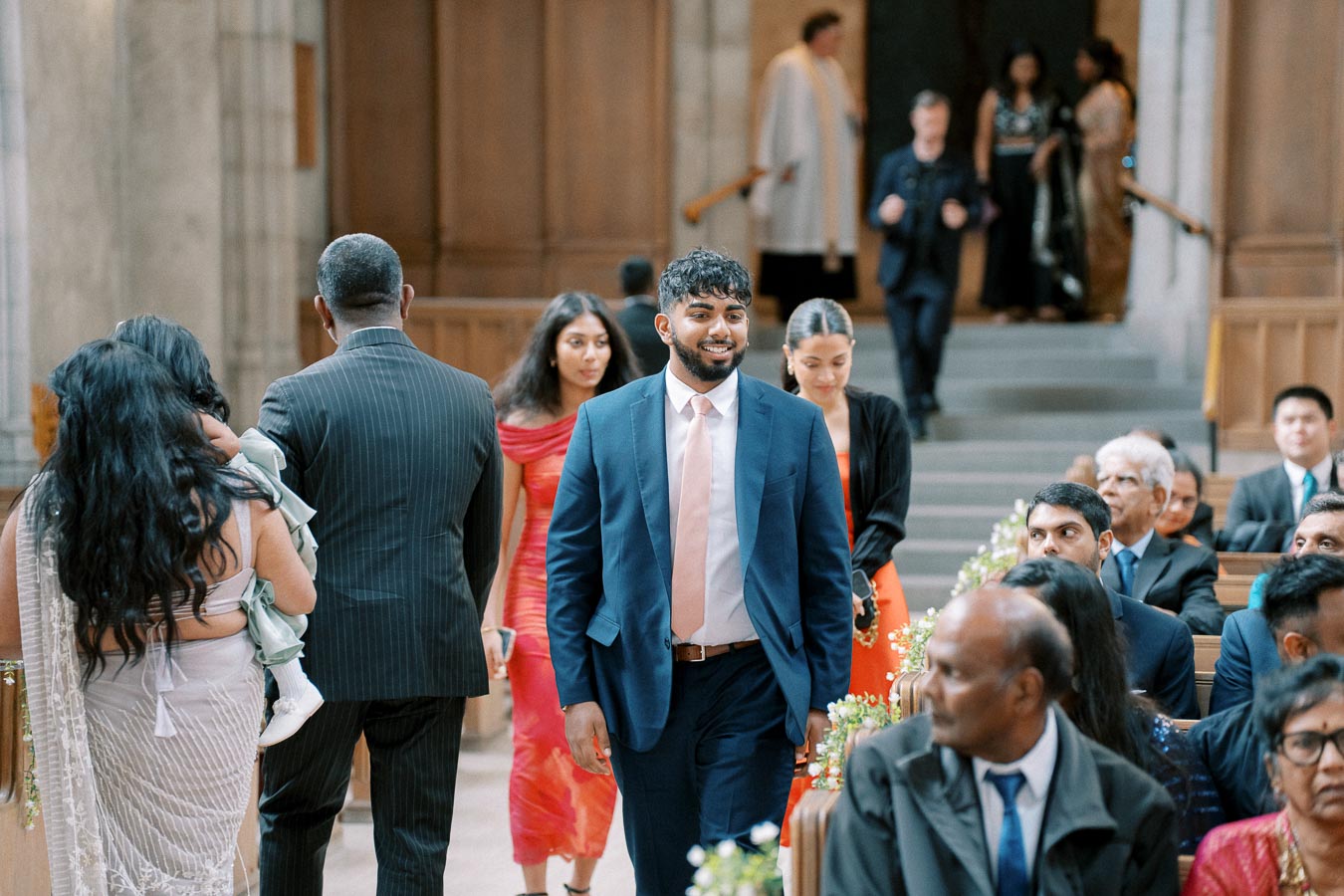 A diverse group of guests dressed in formal attire, walking down the aisle at a wedding ceremony held in an elegant church setting.