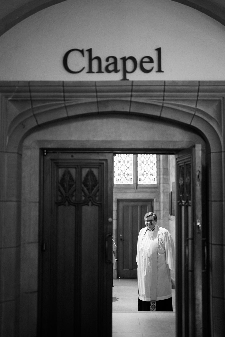 Black and white photograph of a clergy member standing in the entrance of a chapel with arched doorway and stained glass window in the background.