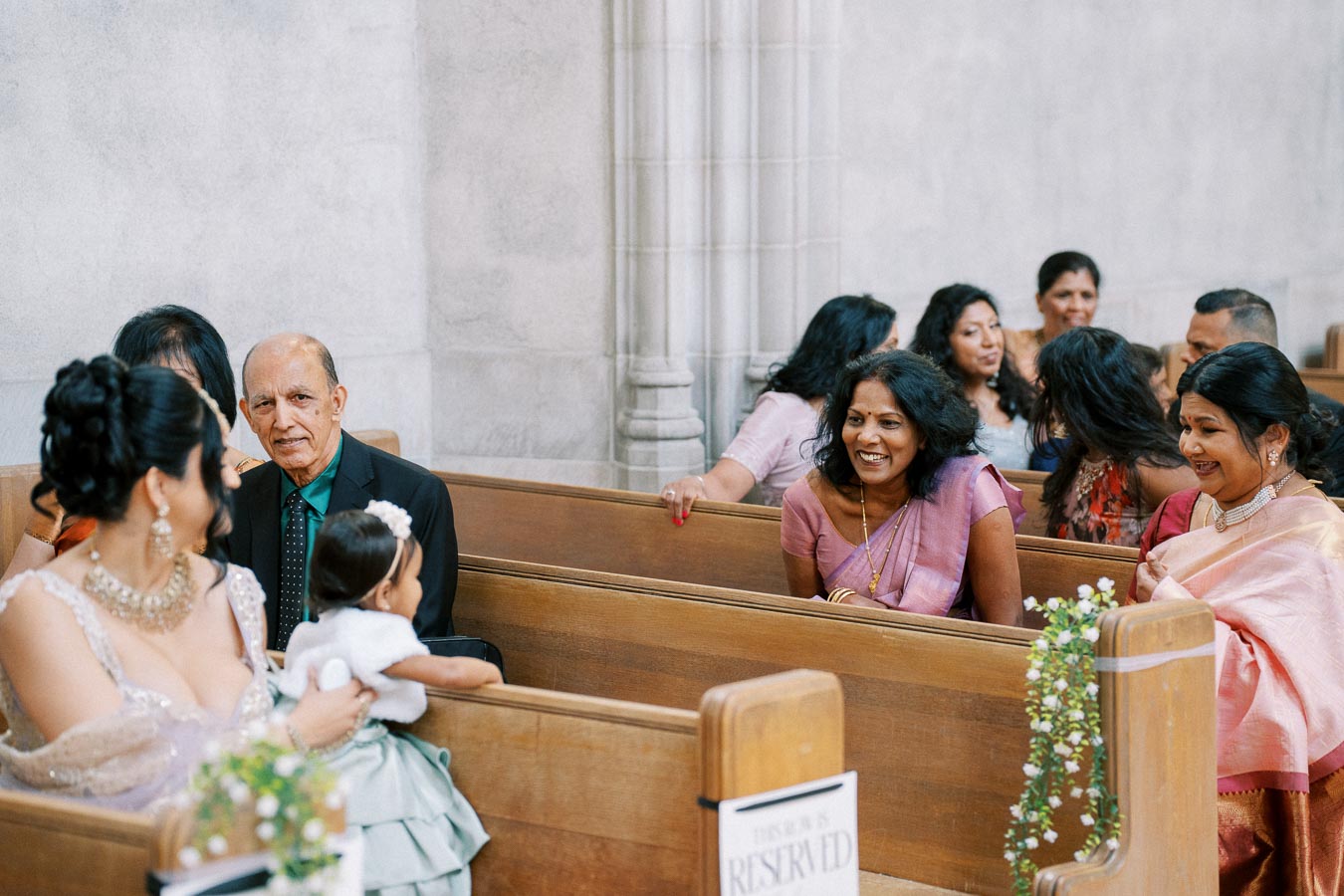 Guests seated in a decorated church pew, engaging and smiling during a wedding ceremony.