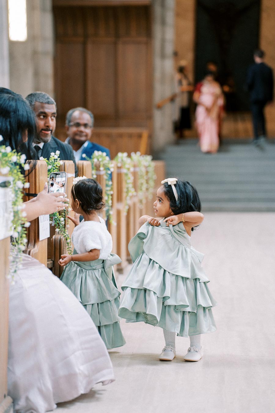 Two young flower girls in matching light green dresses and accessories stand near church pews decorated with flowers, interacting with a person holding a smartphone, while guests observe the scene in the background.