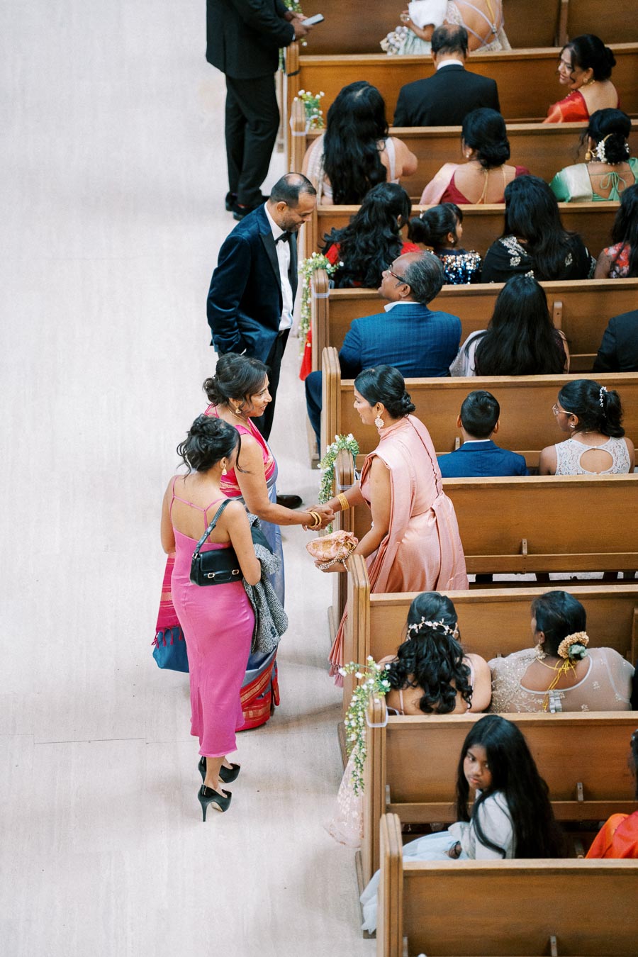 Overhead view of elegantly dressed guests seated in wooden pews at a wedding ceremony. Women in colorful sarees and dresses are seen interacting, with floral decorations on the pews adding a festive touch.