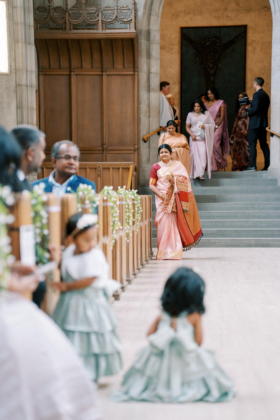A woman in a colorful saree walks down the aisle of a decorated church during a wedding ceremony, with guests observing and children in the foreground.