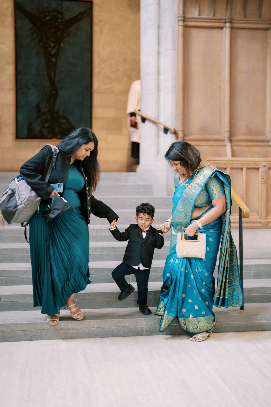 A young boy in a suit holds hands with two women while descending steps at an elegant venue. One woman wears a traditional blue and gold saree, and the other is dressed in a teal gown with a coat. The background features ornate architecture and artwork.