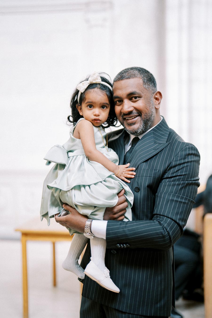 A smiling man in a pinstripe suit holding a young girl in a light blue dress with a bow, standing indoors.