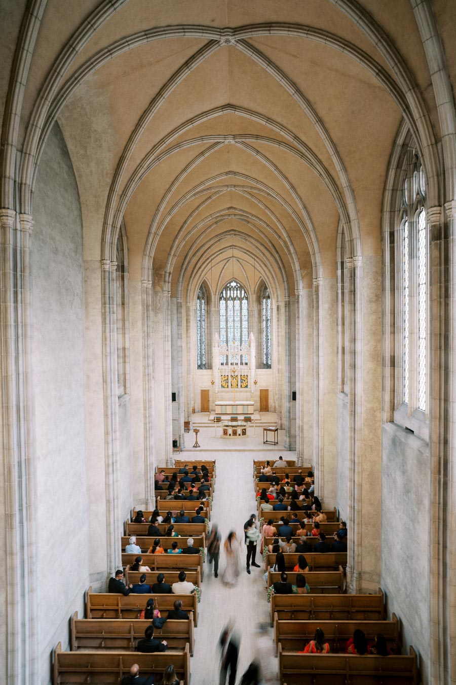 A spacious church interior with high arched ceilings, featuring wooden pews filled with people, leading to a central altar with large stained glass windows in the background.