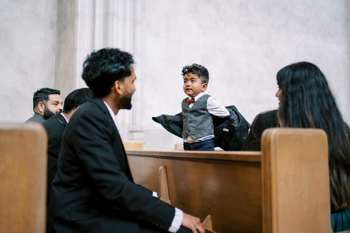 A young boy dressed in formal attire with a bow tie and vest stands proudly in a church pew, surrounded by adults in suits, enjoying a special event or ceremony.