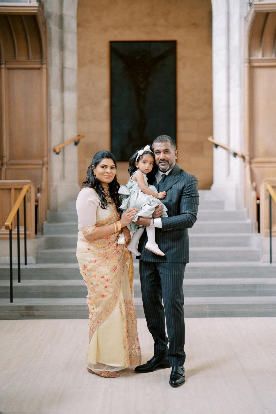A family posing together in an elegant setting with wooden accents and stone stairs. The mother is wearing a traditional saree, the father is in a formal suit, and they are holding a young child dressed in a light-colored dress with a bow.