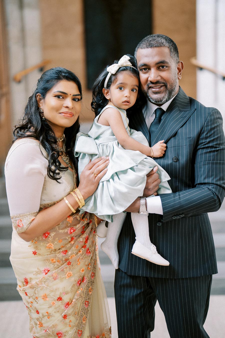 A family portrait with a woman in a traditional saree standing next to a man in a suit, holding a toddler in a light blue dress, capturing a moment of togetherness and elegance.