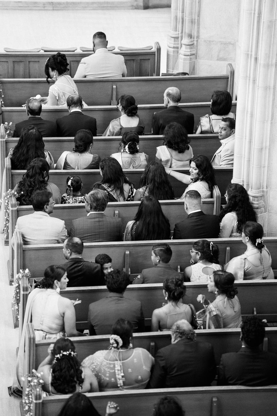 Aerial view of a wedding ceremony in a church, showing guests seated in pews, with elegant attire and floral decorations. Black and white photograph capturing a moment of anticipation.