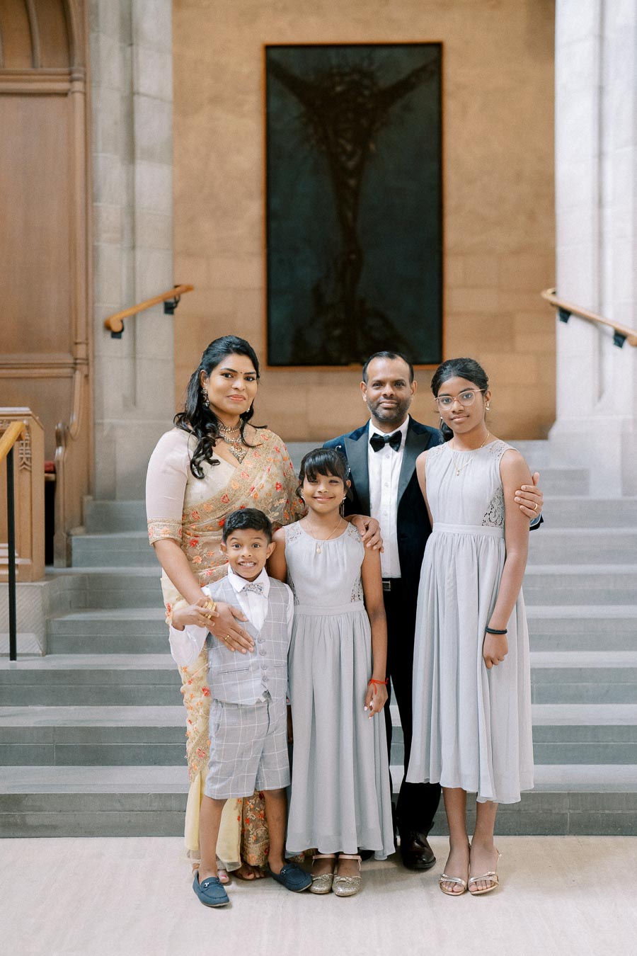 Family posing together in elegant attire on a staircase, with a woman in a saree and three children in formal outfits, set against a sophisticated interior background.