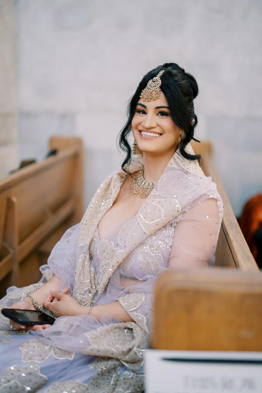 Smiling woman in an elegant traditional outfit sitting in a church pew, adorned with intricate jewelry and holding a phone, blending modern technology with cultural attire.