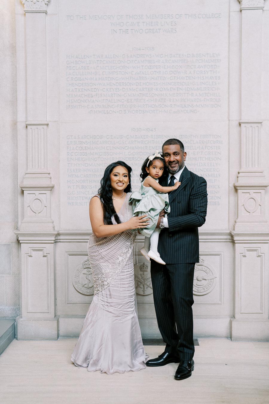 A family dressed elegantly stands together in front of a stone memorial wall. The woman wears a light-colored gown, the man is in a dark suit, and they hold a young child in a green dress. The wall behind them has engraved names as a tribute.