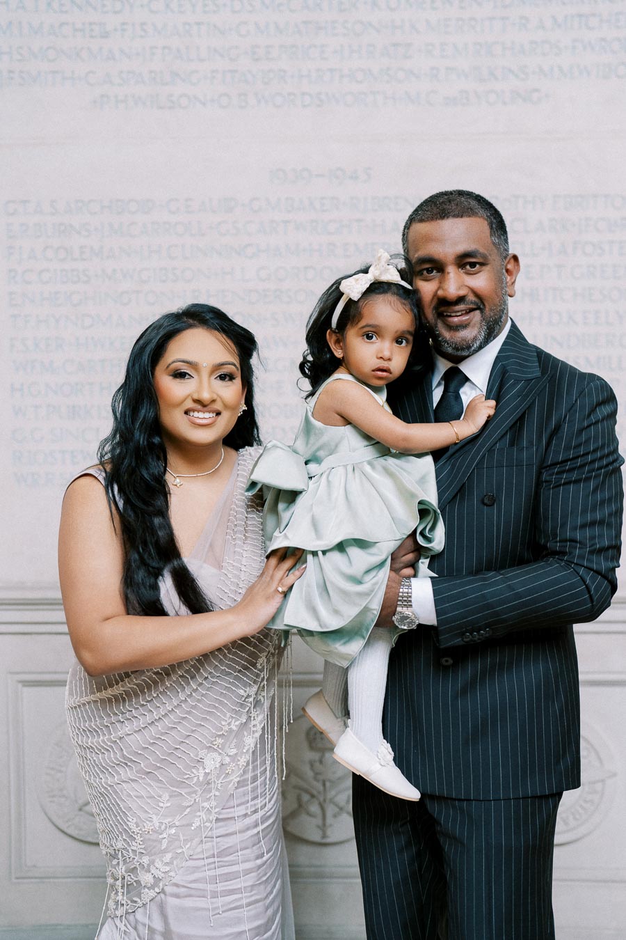 Family dressed in formal attire, featuring a woman in an elegant sari, a man in a pinstripe suit, and a young girl in a green dress, standing against a stone wall backdrop with engraved text.