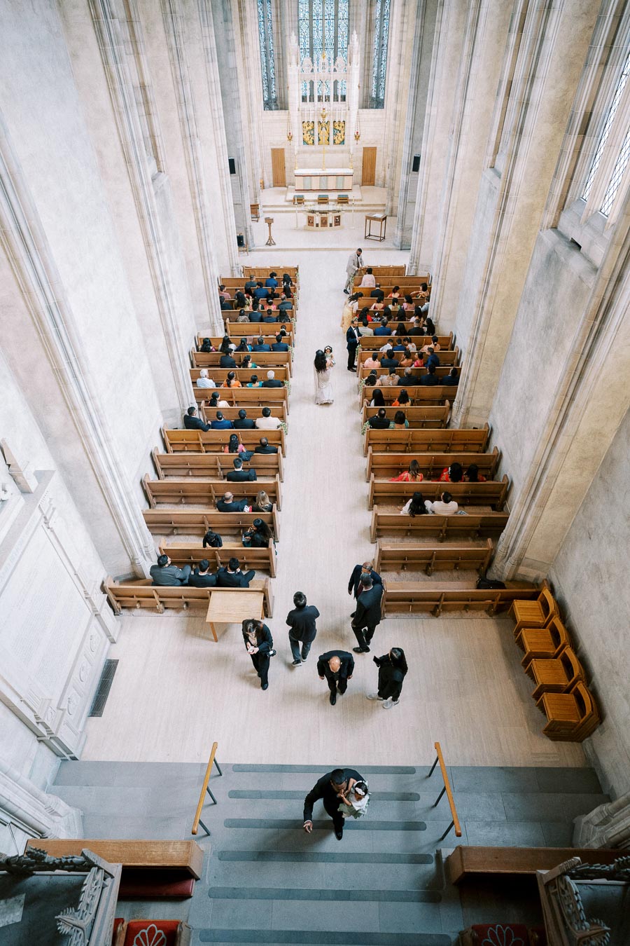 Overhead view of a large, elegant church interior filled with pews and people, featuring a prominent altar and high ceilings with stained glass windows, creating a serene and spiritual atmosphere.