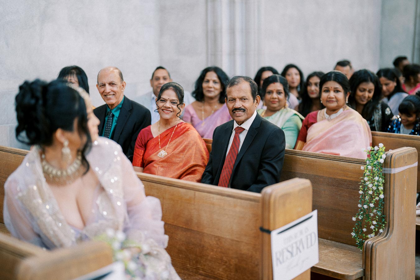 Wedding guests seated in pews, dressed in formal attire, smiling and engaged in conversation. The focus is on a man in a suit and a woman in a red dress, with a blurred woman in traditional attire in the foreground. The setting is decorated with floral elements.