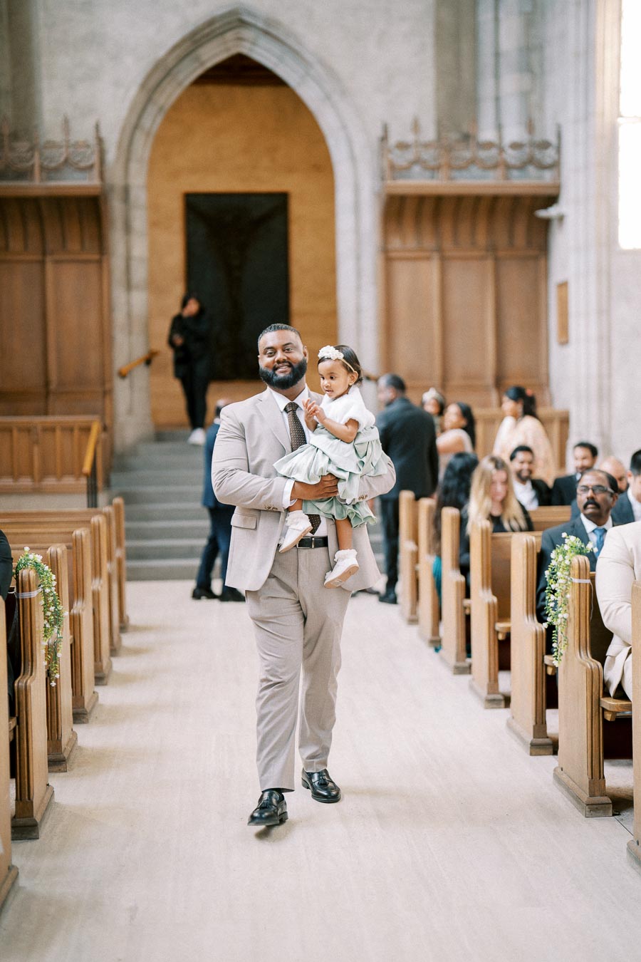 Father in a suit walking down the aisle of a church carrying his young daughter in a formal dress during a wedding ceremony.