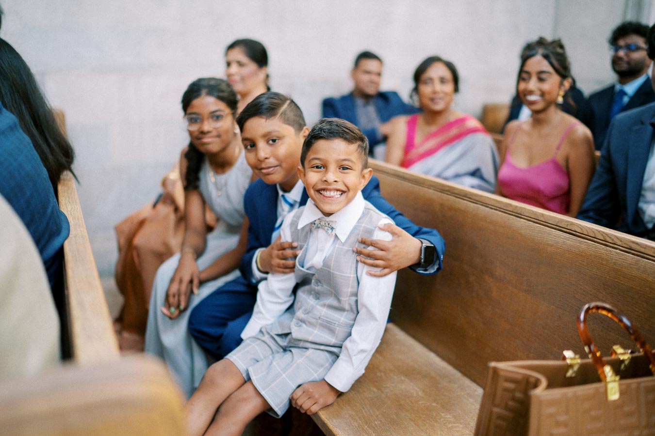 A joyful group of children and adults sitting in a wooden pew, dressed in formal attire, capturing a happy moment at a special event.