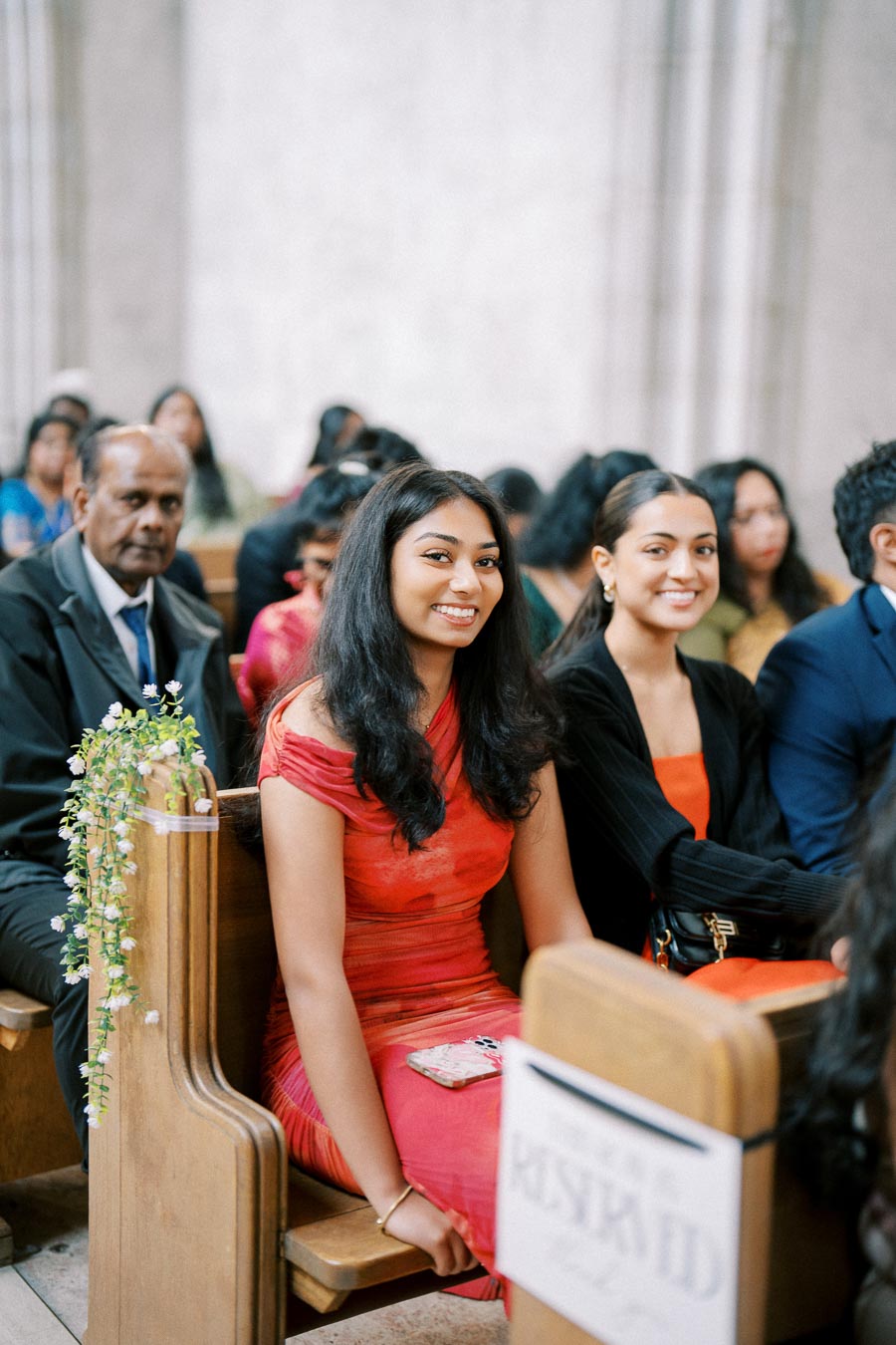 Smiling young women in vibrant attire sitting in a church pew during a formal event, surrounded by other attendees, with floral decorations on the bench.