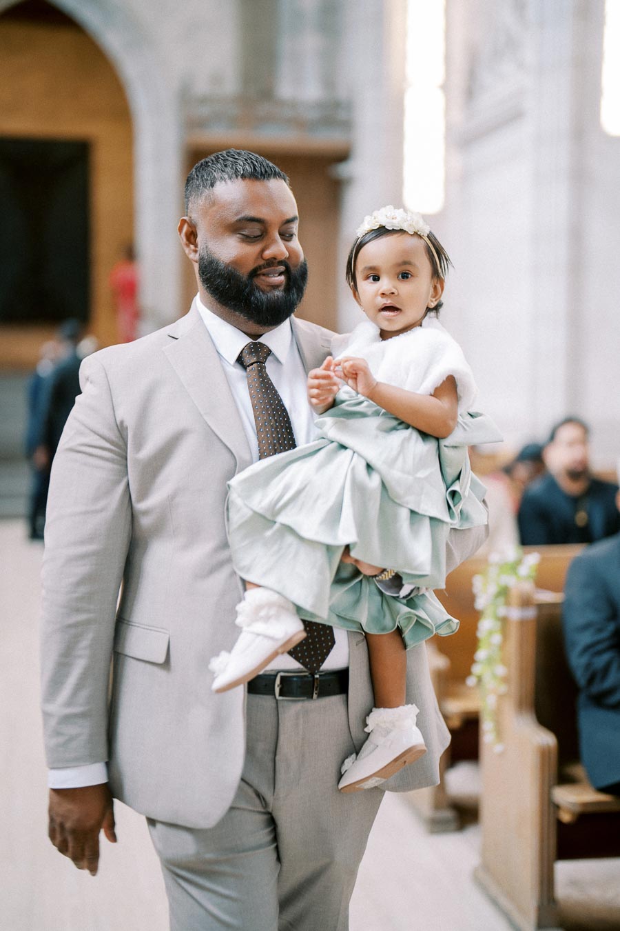 Father in a light gray suit carrying his young daughter wearing a pastel dress and floral headband, walking down an aisle in a beautifully lit church.