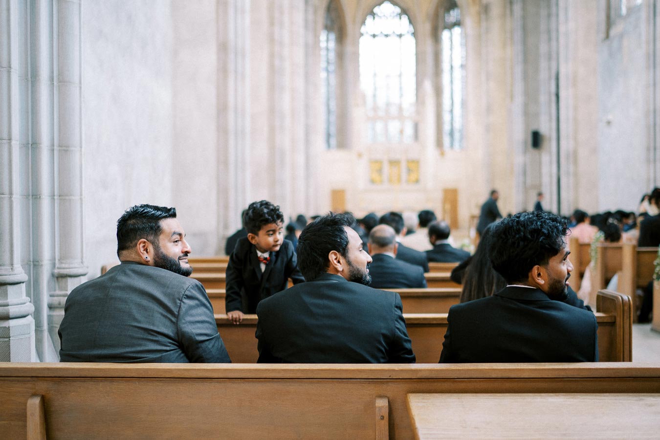 Formal gathering in a spacious church interior, featuring elegantly dressed men seated on wooden pews, with one looking back and a child peering over the seat. Sunlight filters through large stained glass windows in the background.