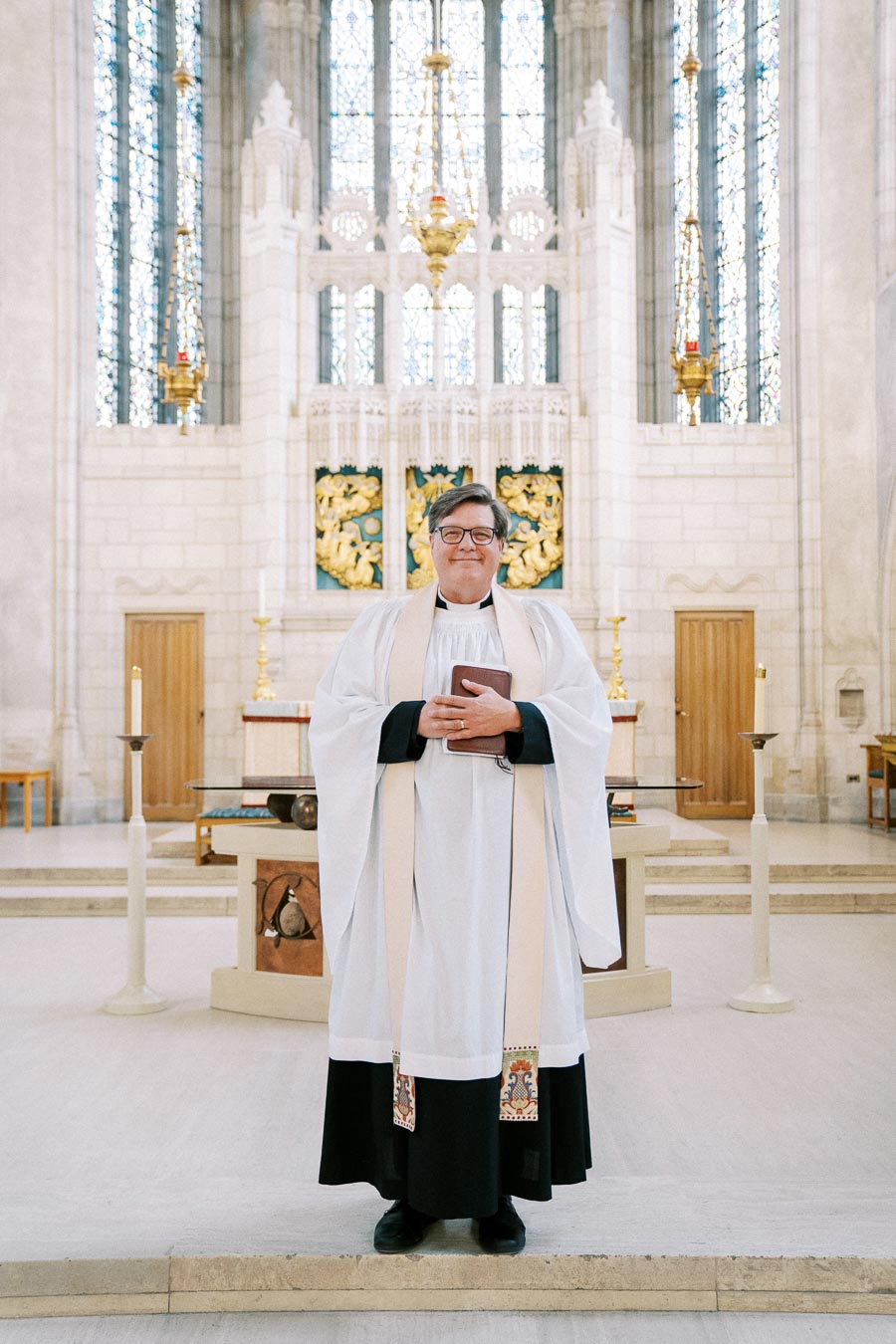 A person in a white clerical robe stands inside a grand church, holding a book. The background features intricate stained glass windows and ornamental gold details, creating a serene and spiritual atmosphere.