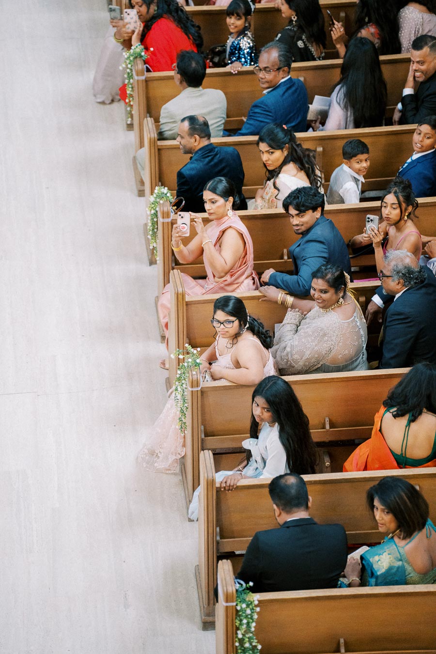 Guests seated in a beautifully decorated church pew, dressed in formal attire, capturing moments on their phones during a wedding ceremony.