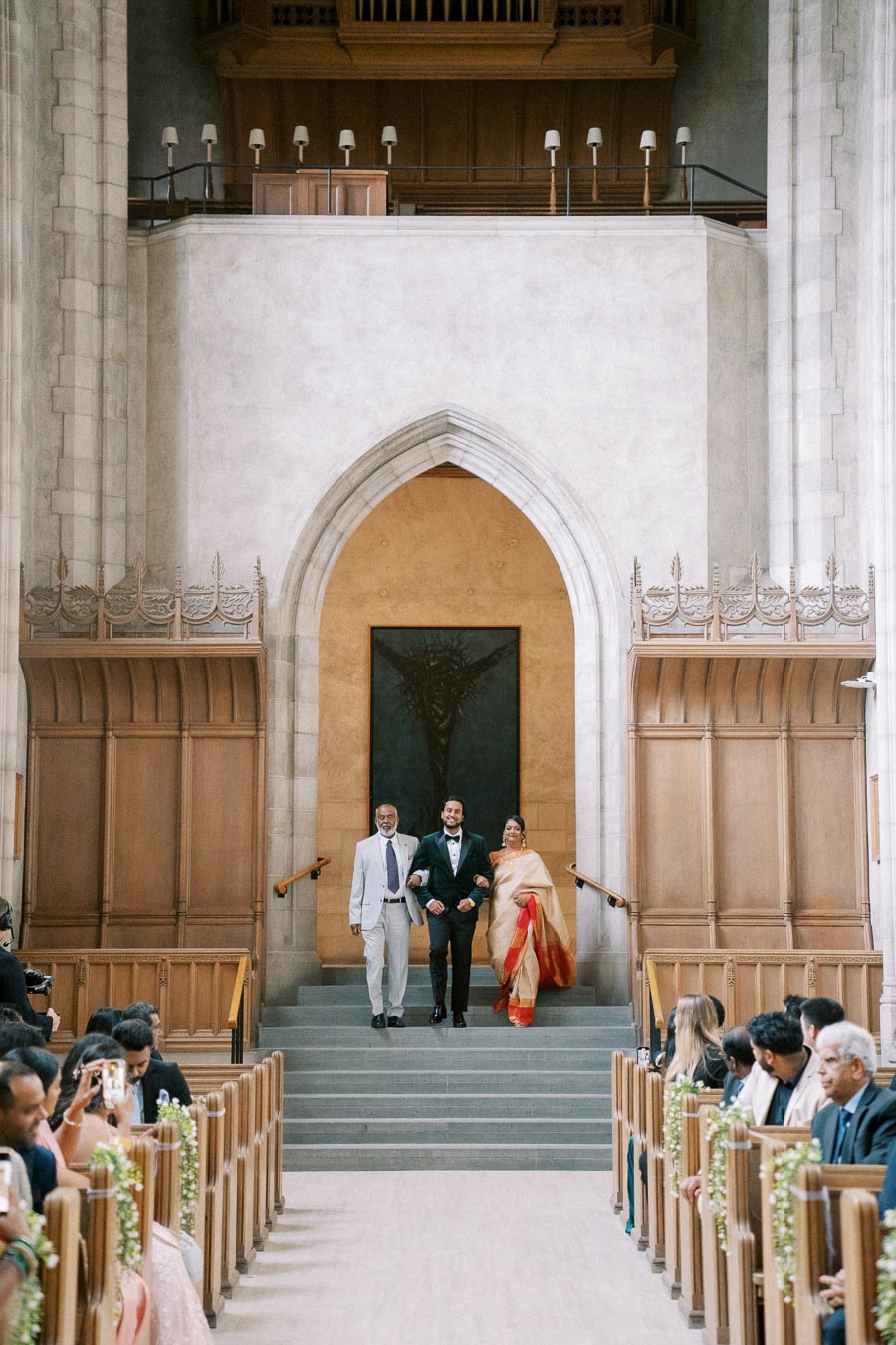 A groom walking down the aisle with two family members, one wearing a white suit and the other in a traditional saree, in a beautifully adorned chapel with guests seated and capturing the moment.