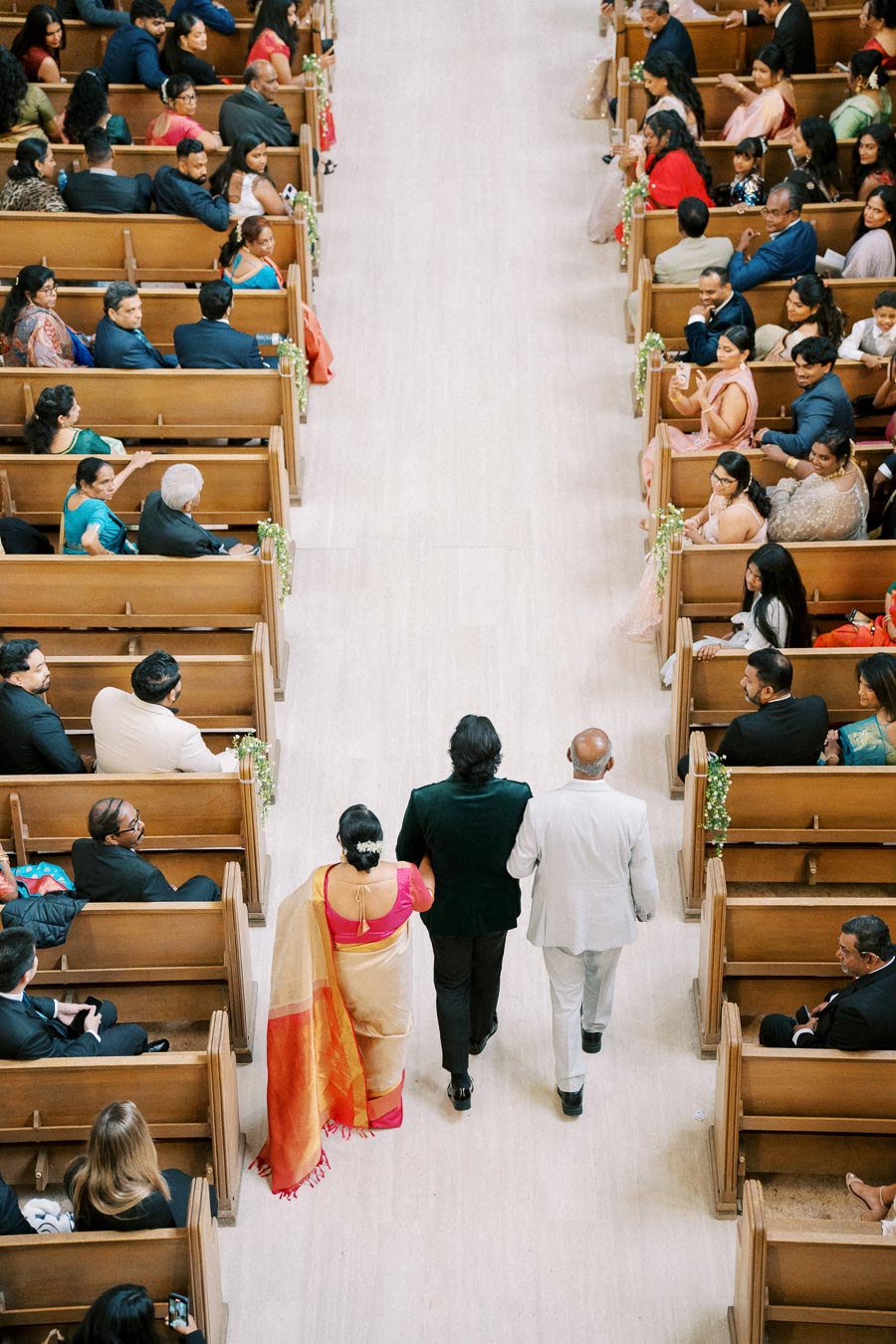 A trio walking down the aisle in a wedding ceremony venue, surrounded by seated guests on wooden pews, with vibrant attire and elegant decorations creating a festive atmosphere.