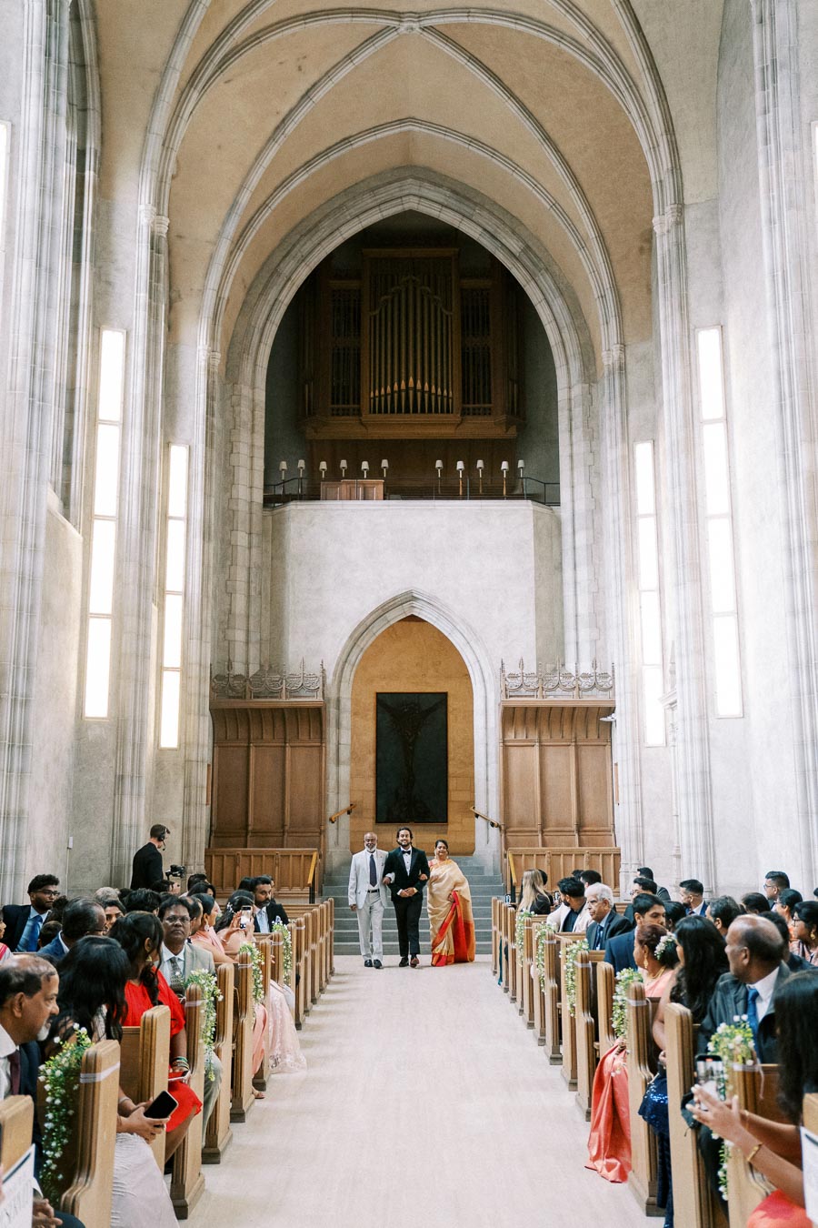 Elegant wedding ceremony inside a grand cathedral with high vaulted ceilings, featuring guests seated on both sides of the aisle decorated with flowers, as the groom walks down the aisle with his parents.