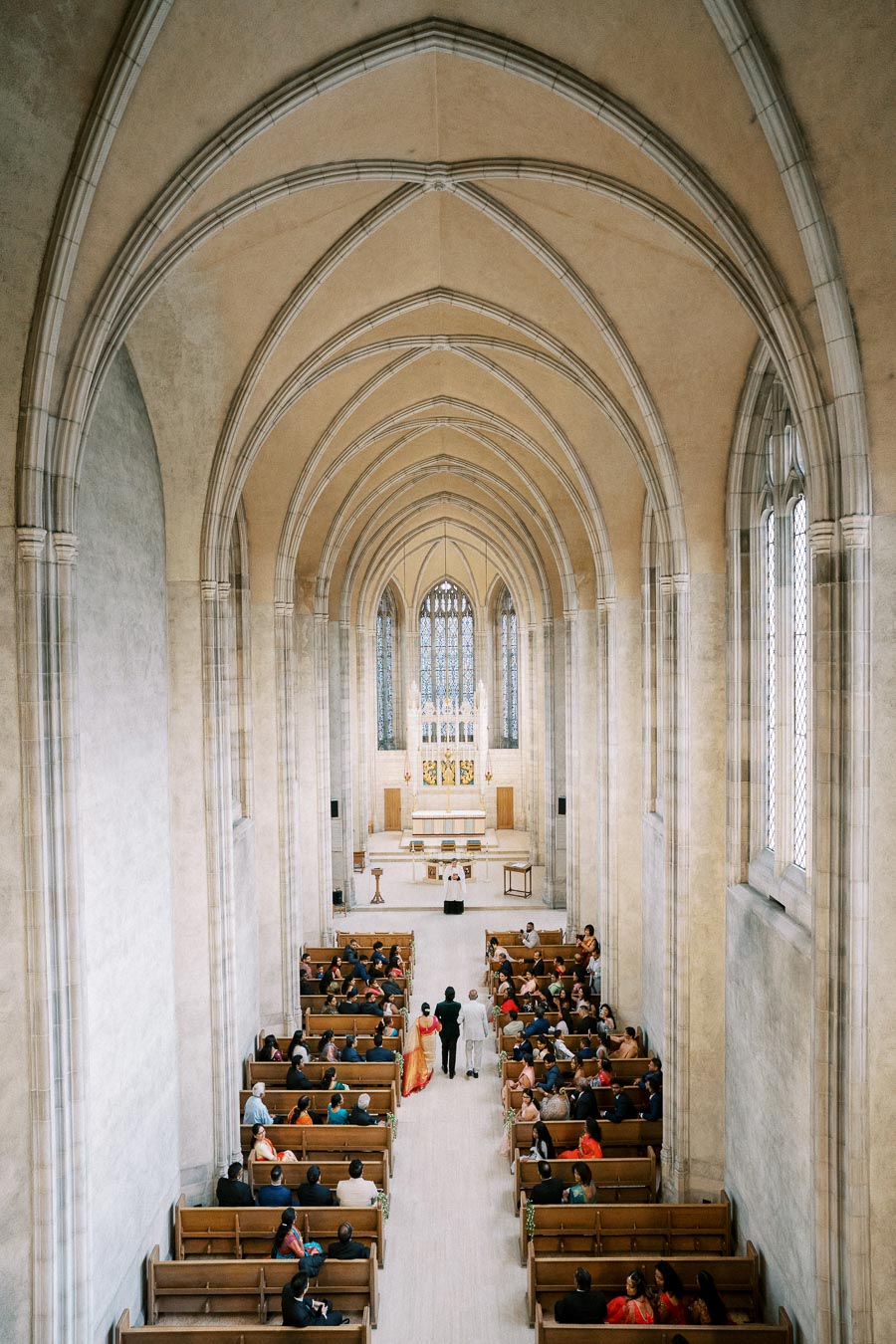 A grand cathedral interior with a high vaulted ceiling and stained glass windows, featuring a wedding ceremony in progress with guests seated in pews and a couple approaching the altar.