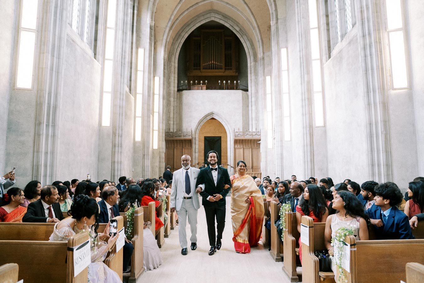 A groom in a black tuxedo walks down the aisle with his parents in a grand, light-filled church, surrounded by seated guests during a wedding ceremony.