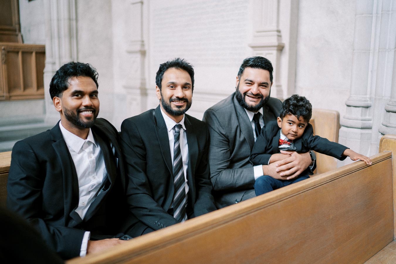 Three men in formal attire sitting on a wooden bench with a young boy in a suit, smiling inside a bright room with stone walls.