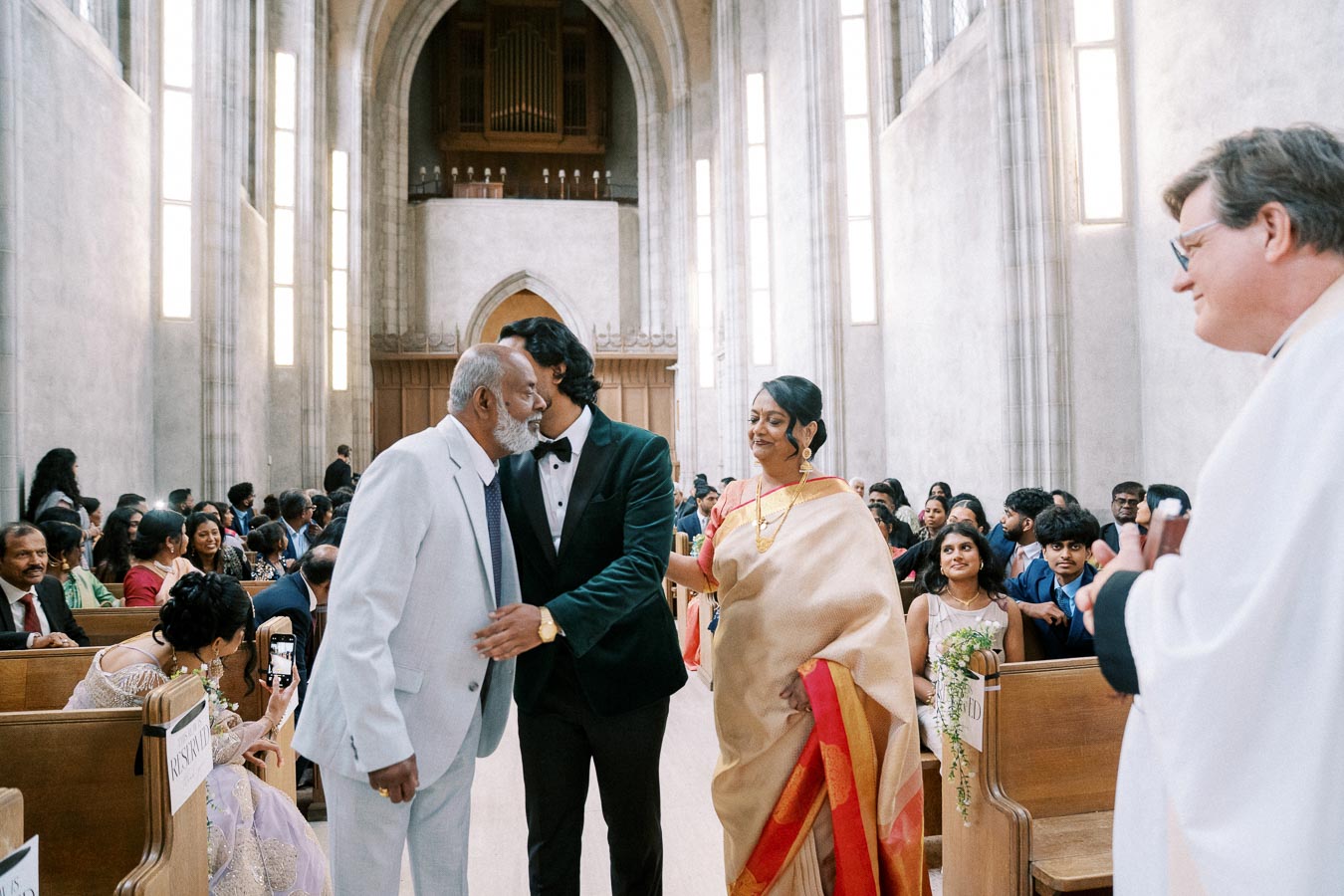 Ceremony in a church with a man in a tuxedo greeting an elderly man in a white suit, accompanied by a woman in a traditional saree, surrounded by seated guests.