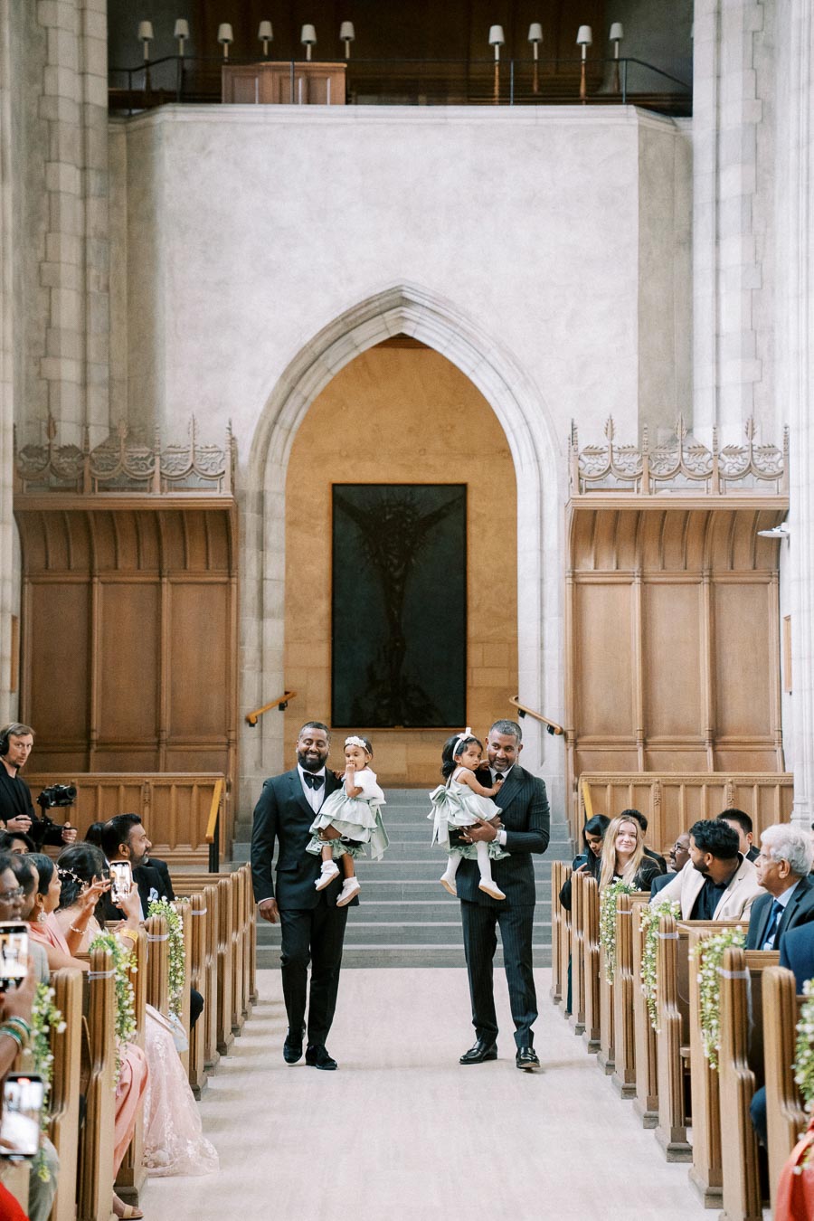 A joyful wedding ceremony inside an elegant church, featuring two men in suits walking down the aisle, each holding a young child. The guests, seated on wooden benches adorned with greenery, capture the moment on their phones. The church is beautifully decorated with tall archways and ornate details.
