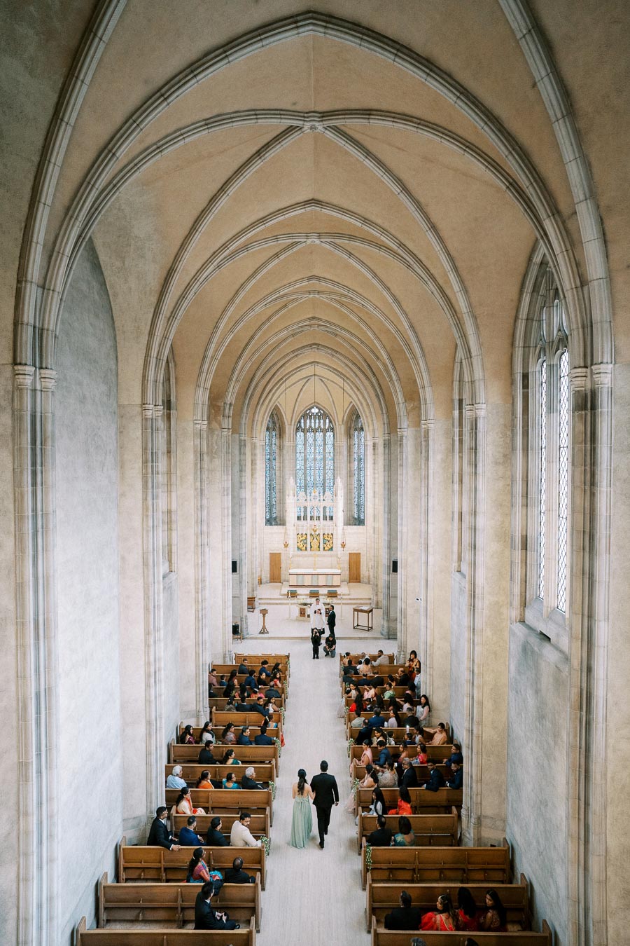 Interior of a grand church with arched ceilings and stained glass windows, featuring a wedding ceremony with guests seated and a couple walking down the aisle.