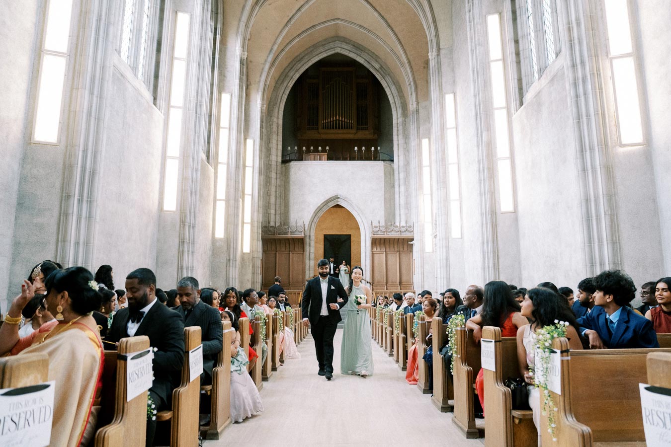 Elegant church wedding ceremony with a couple walking down the aisle, surrounded by seated guests in formal attire, in a beautifully lit cathedral with high arches and stained glass windows.