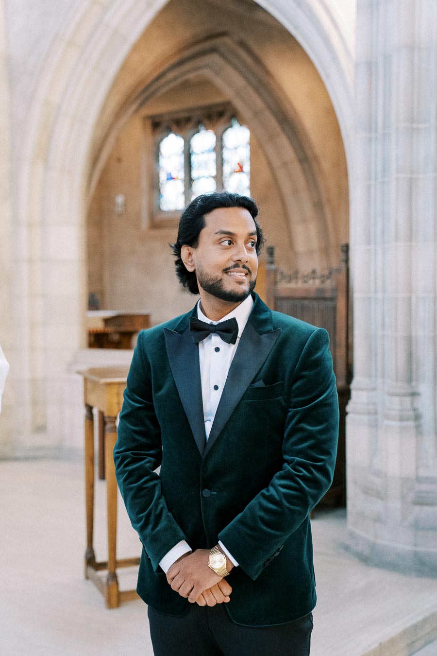 A well-dressed man in a formal teal velvet suit with a black bow tie, standing inside a church with arched architecture and a stained glass window in the background.