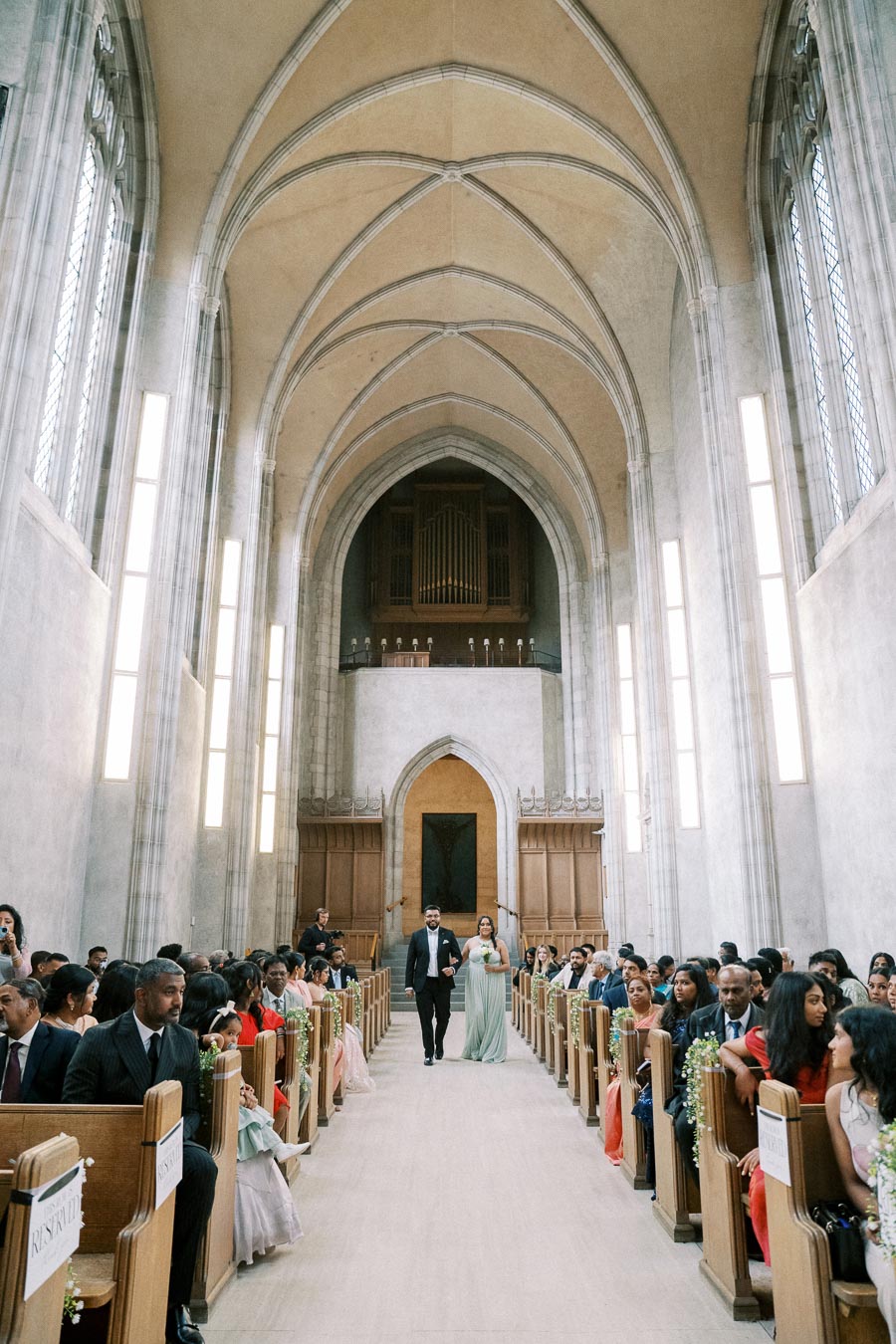 Wedding ceremony in a grand cathedral with ornate arches, guests seated in wooden pews, and a wedding party member walking down the aisle holding a bouquet.
