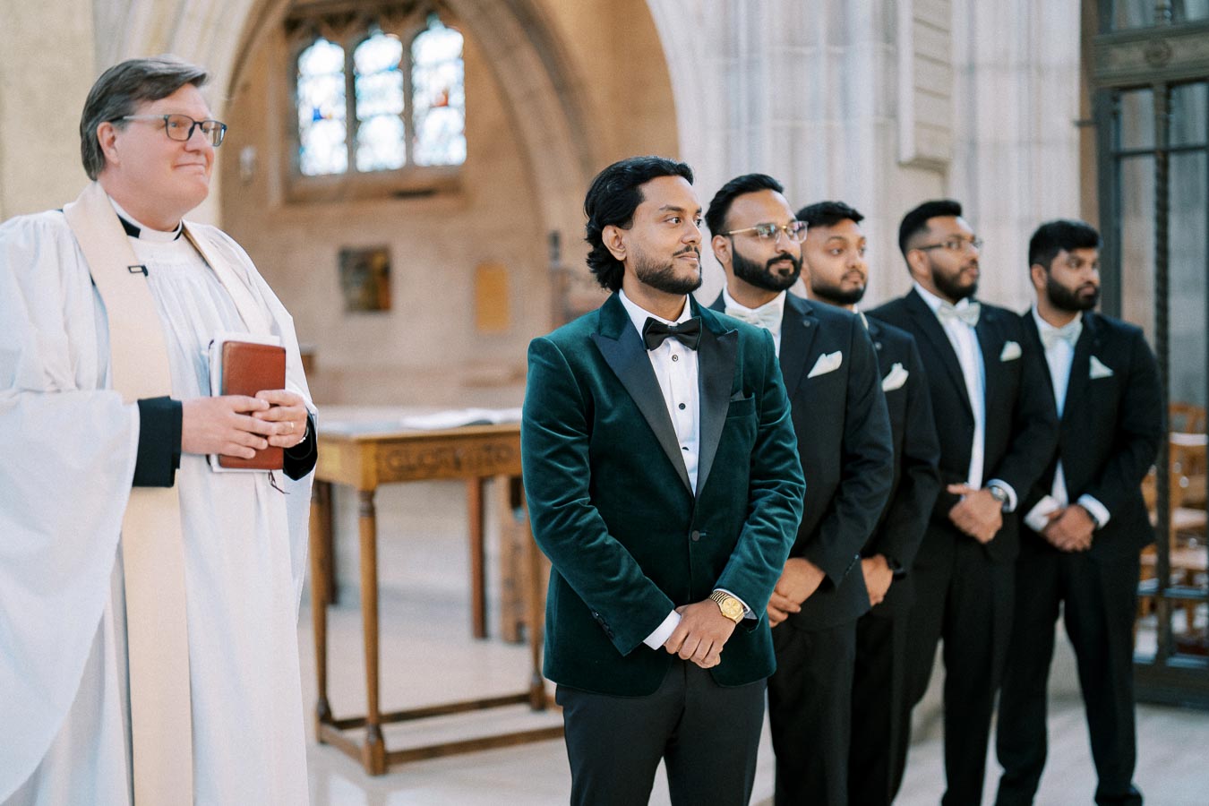 Groom in elegant green velvet tuxedo with groomsmen and officiant in a church setting, awaiting wedding ceremony.