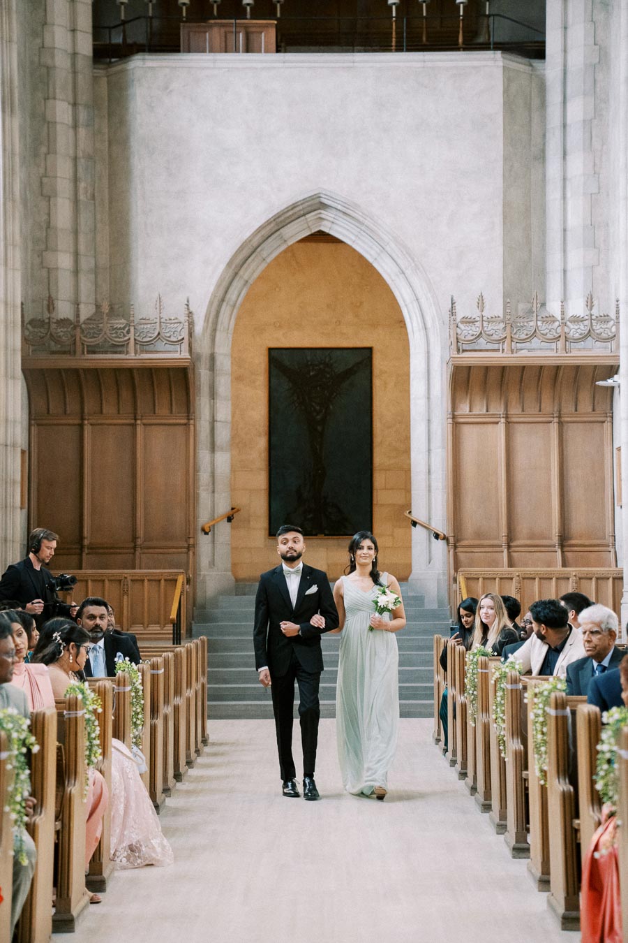 Wedding ceremony in a grand church, featuring a couple walking down the aisle. The man wears a black suit, while the woman is in a light-colored dress holding flowers. Guests are seated on wooden pews adorned with floral decorations, with a large archway and artwork in the background.